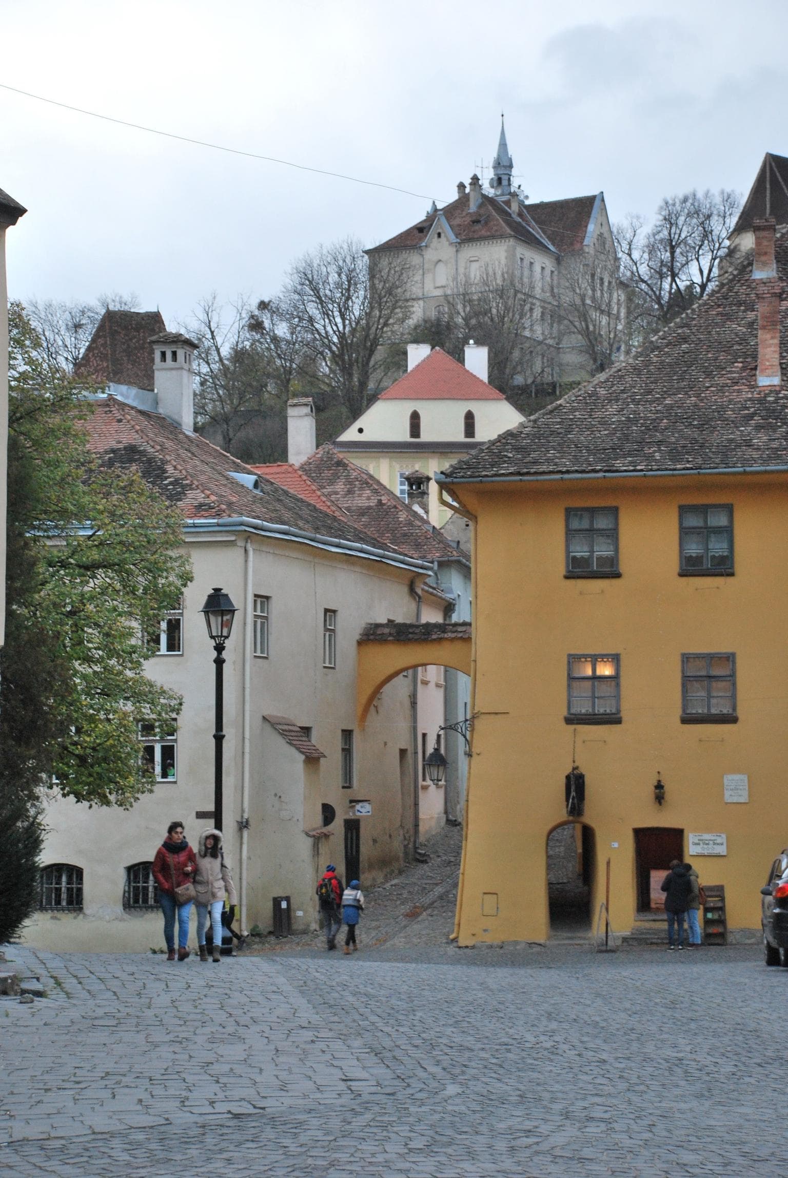 Narrow cobblestone street with yellow buildings and pedestrians; medieval castle sits on hill in background.
