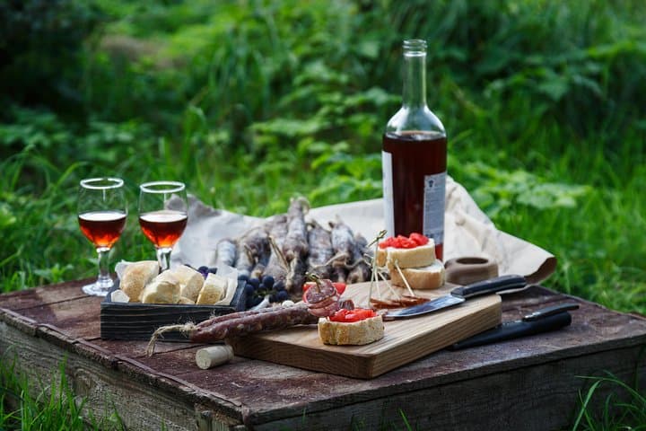 Outdoor picnic scene with wine bottle, two glasses, bread and cured meats on wooden crate.