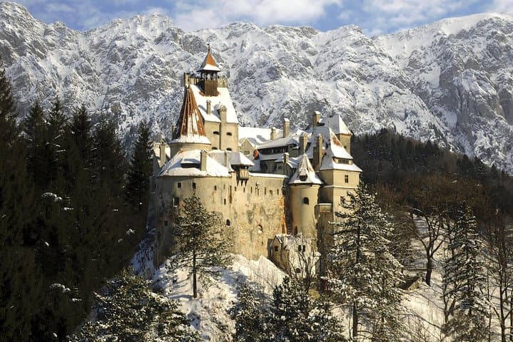Snow-covered medieval castle perched on forested hill with rugged mountains and clear winter sky.