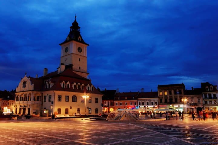 Twilight town square with illuminated clock tower, central fountain, bustling pedestrians, and deep blue sky.