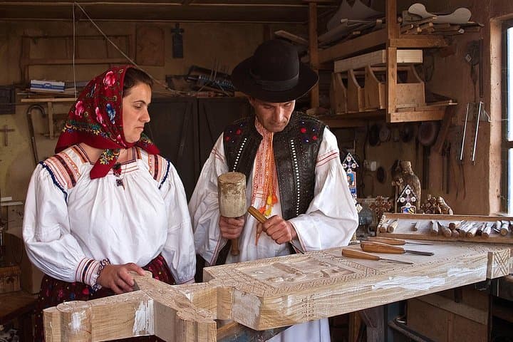 Couple in traditional folk dress carving ornate wood, man chiseling while woman observes in a workshop.